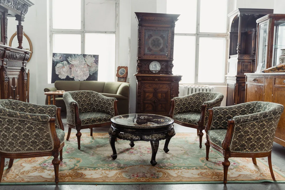 Upholstered antique armchairs around a lacquered table on a rug, with carved cabinets and a tall case clock behind, set out like a sale preview
