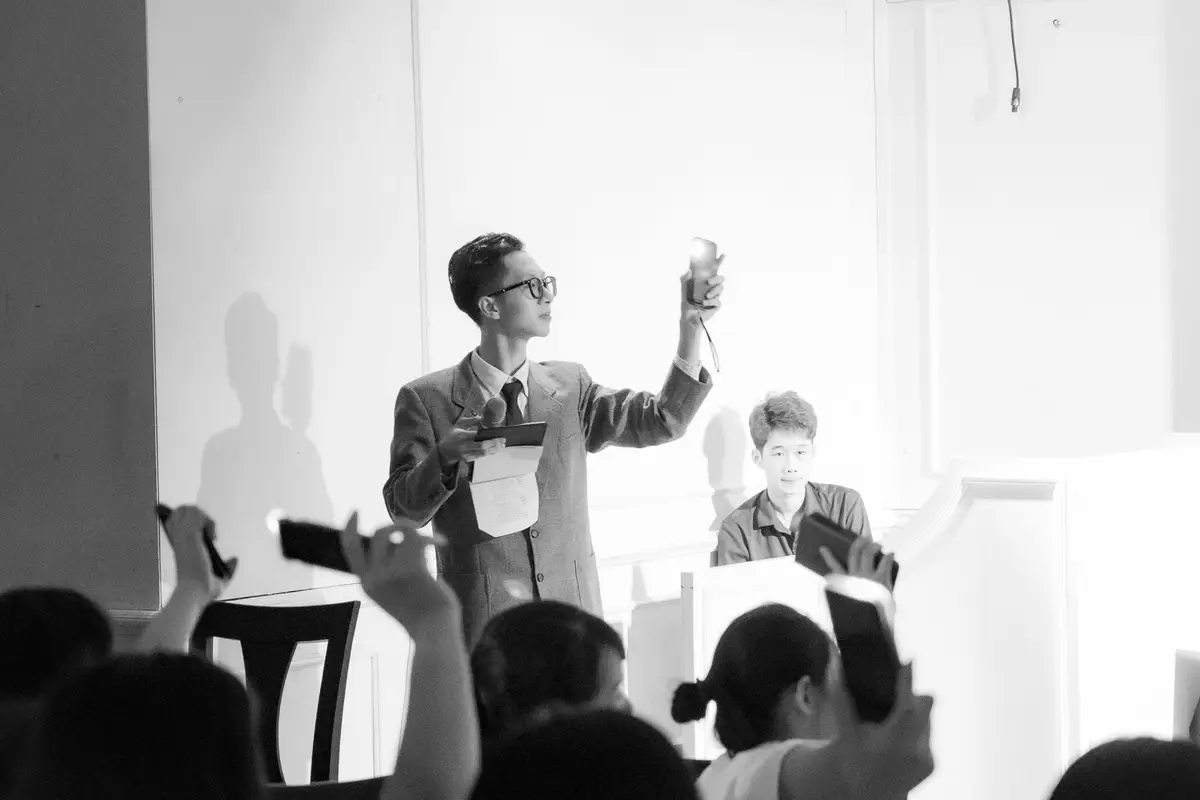 Black and white photo of an auction in progress, with an auctioneer at the front and bidders in the room holding up phones