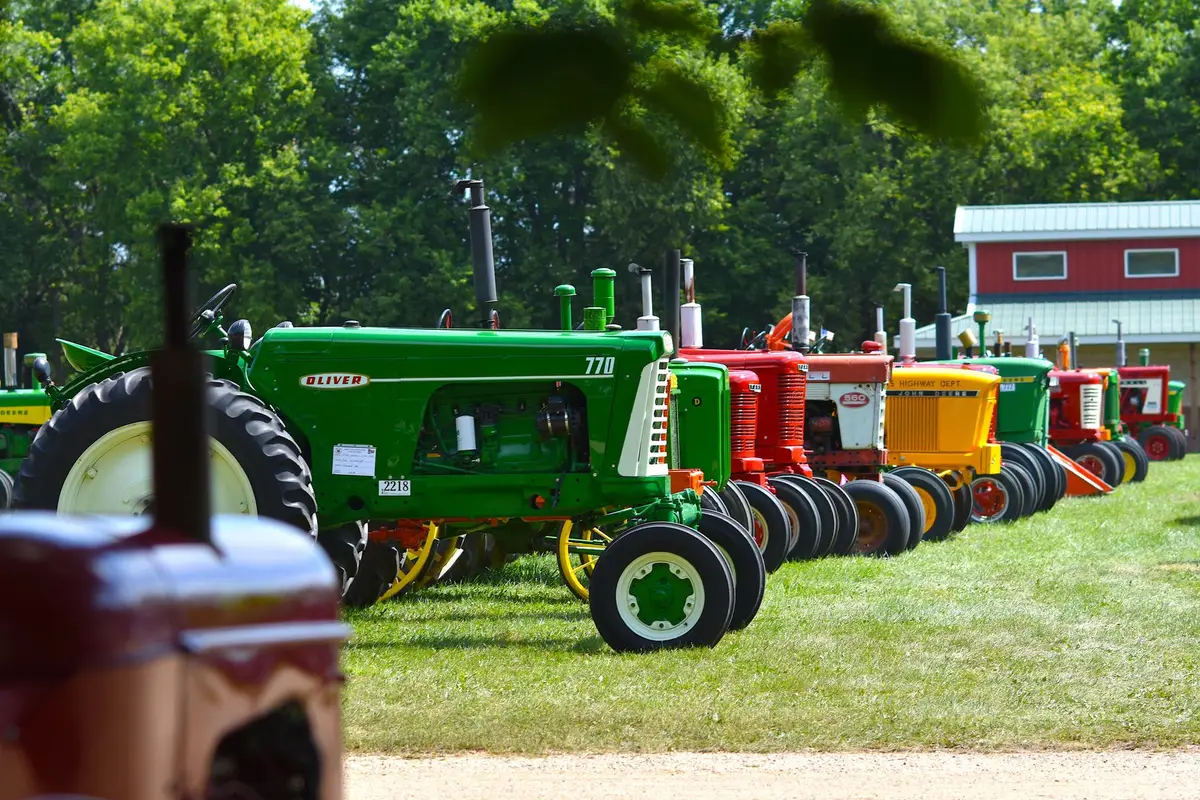 A row of green, red and yellow vintage tractors lined up on grass in front of a red barn, the way equipment is lined up for a farm sale